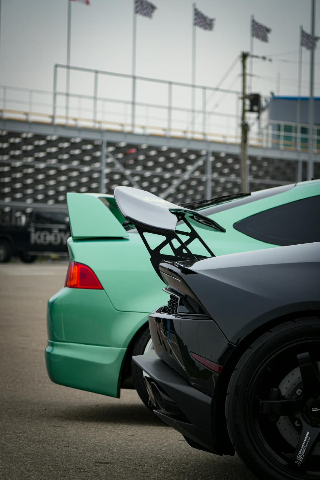 Rear wing and body details of a dark Lamborghini beside a mint-green car at a racetrack
